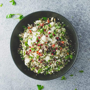 A round black bowl with a mix of cauliflower florets, roasted root vegetables, hazelnuts, toasted spicy chickpeas, cranberries, and mixed herbs, with a small bowl of lemon vinaigrette on a marble table.