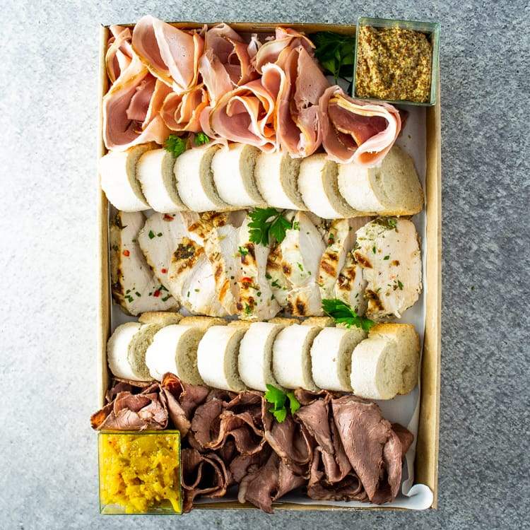 A large rectangular cardboard box containing slices of roast beef, ham, and chicken breast, with mustard, chutney, and sliced baguette.