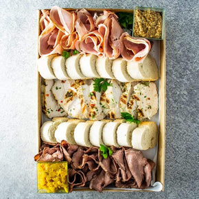 A large rectangular cardboard box containing slices of roast beef, ham, and chicken breast, with mustard, chutney, and sliced baguette.