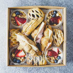 A box of assorted pastries and breakfast pots including croissants, fruit danish, and granola yogurt tubs.