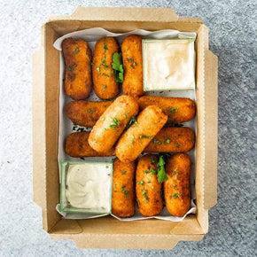 A rectangular cardboard box containing 12 golden croquettes including flavours like salt cod and potato or truffle and potato, with tubs of dipping sauces, and parsley garnish on a marble table.