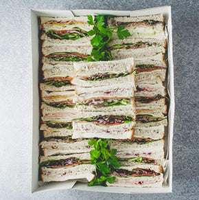 A large rectangular cardboard box containing a variety of finger sandwiches, garnished with parsley, on a marble table.