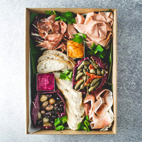 A large rectangular cardboard box containing antipasto, including salami, prosciutto, mortadella, olives, dips, pickled vegetables, and lavosh, on a marble table.