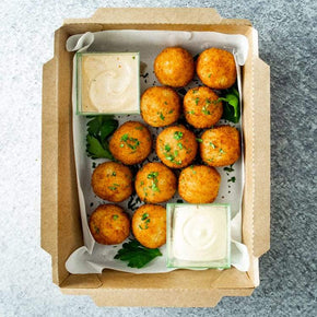 A rectangular cardboard box containing 12 golden arancini, tubs of dipping sauces, and parsley garnish on a marble table.