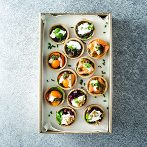 A rectangular cardboard box with a mix of mini savoury tarts including flavours like hot smoked salmon, beetroot, cherry tomato and caramelised onion, on a marble table.
