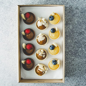 A rectangular cardboard box containing 12 sweet mini tarts with flavours including lemon curd, chocolate, and caramel, on a marble table.