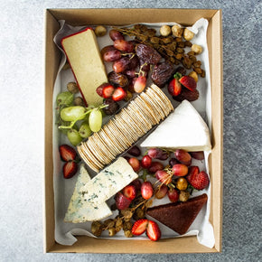 A large rectangular cardboard box containing wedges of 
Australian and European cheeses, quince paste, fresh & dried fruits, and crackers on a marble table.
