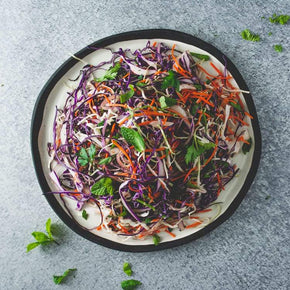 A large round white platter with a mix of red and white cabbage, carrot, onion, mint, and parsley, with a small bowl of buttermilk dressing on a marble table.