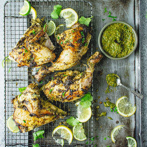 A rectangular metal baking tray with four pieces of Chimichurri chicken, surrounded by slices of lemon, lime, and coriander, with a bowl and spoon of Chimichurri sauce at the side.