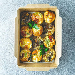 A rectangular cardboard box with 12 mini frittatas with fillings like ham and asparagus, pancetta and tomato, mushroom and cheese, or pumpkin and feta, on a marble table.