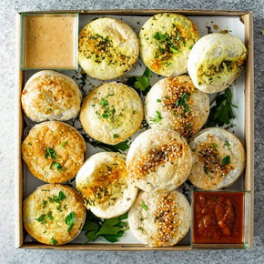 A square cardboard box containing 12 mini pies with fillings like beef, chicken or mushroom, with tubs of dipping sauces on a marble table.