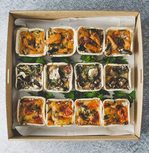 A square cardboard box containing 12 mini quiches in flavours like lorraine, mushroom, vegetable, leek, or smoked salmon on a marble table.