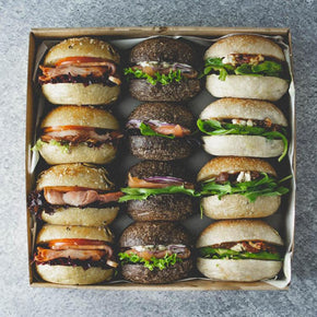 A square box of 12 mini breakfast rolls with fillings like avocado, haloumi, bacon, and smoked salmon on a marble table.