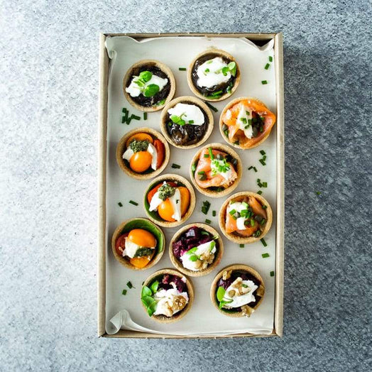 A rectangular cardboard box with a mix of mini savoury tarts including flavours like hot smoked salmon, beetroot, cherry tomato and caramelised onion, on a marble table.