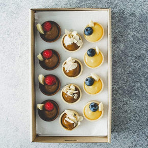 A rectangular cardboard box containing 12 sweet mini tarts with flavours including lemon curd, chocolate, and caramel, on a marble table.