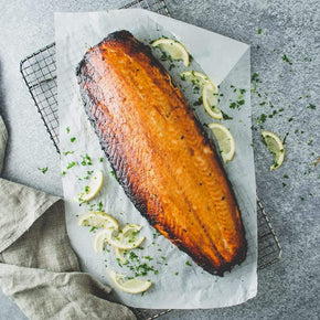 A whole side of baked miso glazed salmon on baking paper, garnished with lemon slices and parsley, on a marble table.