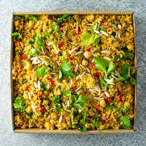 A rectangular cardboard box with a mix of couscous, chickpeas, toasted almonds, currants, capsicums, mixed seeds and herbs, with a tub of lemon vinaigrette on a marble table.