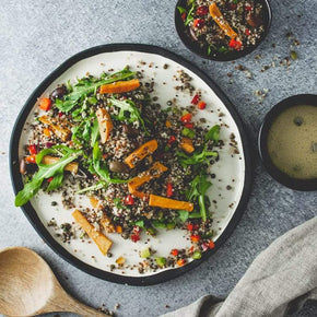 A large round white platter with a mix of French green lentils, quinoa, roasted sweet potato, and mixed seeds, with a small bowl of lemon sumac dressing on a marble table.