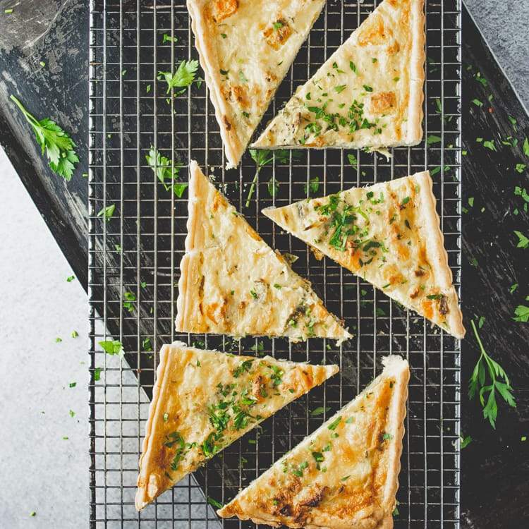 Triangular slices of roast pumpkin, leek, and feta tart on a cooling rack, garnished with parsley, on a marble table.
