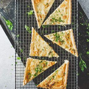 Triangular slices of roast pumpkin, leek, and feta tart on a cooling rack, garnished with parsley, on a marble table.