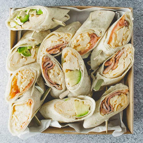 A square box of six savoury breakfast wraps cut in half, with fillings like bacon and egg, salmon and egg, and avocado and egg on a marble table.