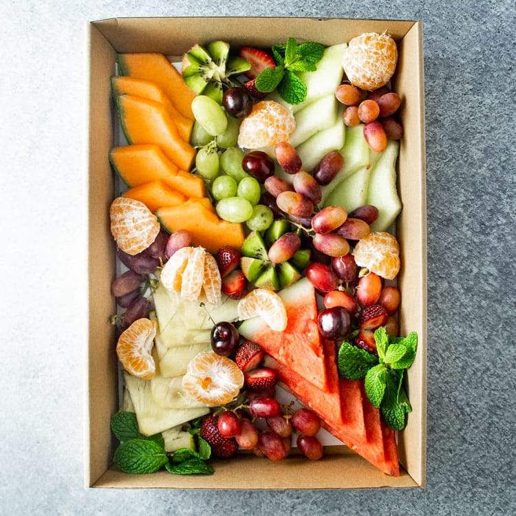 A rectangular box of sliced seasonal fresh fruit including rockmelon, watermelon, grapes, pineapple, kiwi fruit and strawberries on a marble table.