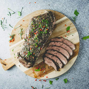 A whole slow cooked, deboned, red wine marinated lamb roast, partially sliced, garnished with herbs, on a wooden board.