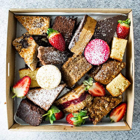 A square cardboard box filled with assorted sweet slices and macarons, dusted in icing sugar with a strawberry garnish on a marble table.