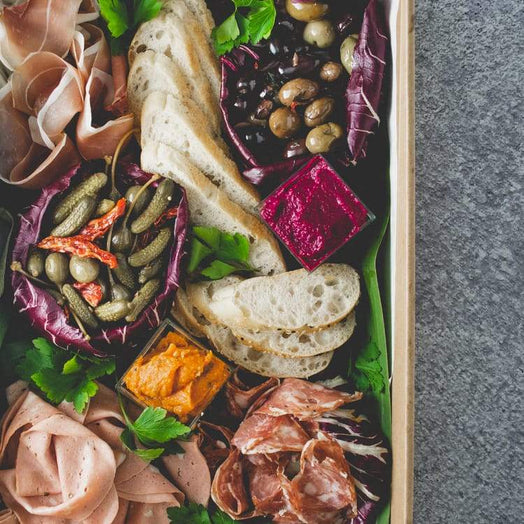 Close up of a large rectangular cardboard box containing antipasto, including salami, prosciutto, mortadella, olives, dips, pickled vegetables, and lavosh, on a marble table.