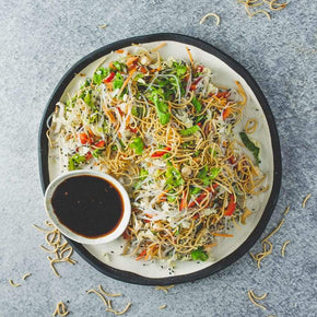 A large round plate with a mix of rice vermicelli, snow peas, mung beans, wombok, capsicum, chilli, and sesame seeds, with a small bowl of lime soy dressing on a marble table.
