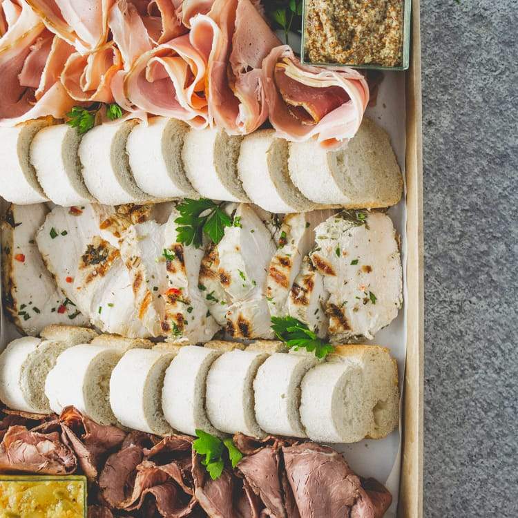 Close up of a large rectangular cardboard box containing slices of roast beef, ham, and chicken breast, with mustard, chutney, and sliced baguette.