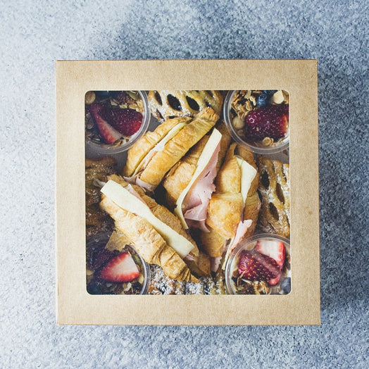 A box with a lid containing assorted pastries and breakfast pots including croissants, fruit danish, and granola yogurt tubs.
