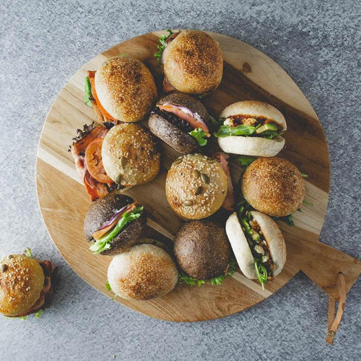A round wooden board with 12 mini breakfast rolls with fillings like avocado, haloumi, bacon, and smoked salmon on a marble table.