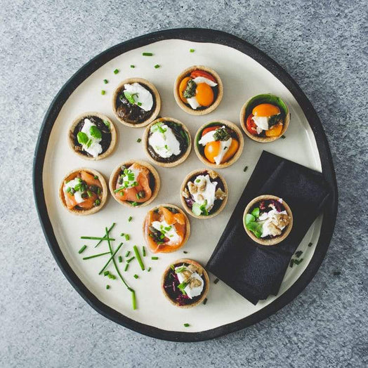 A large round white platter with a mix of mini savoury tarts including flavours like hot smoked salmon, beetroot, cherry tomato and caramelised onion, on a marble table.