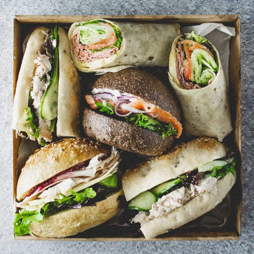 A square cardboard box containing an assortment of rolls and wraps with a mix of fillings, on a marble table.