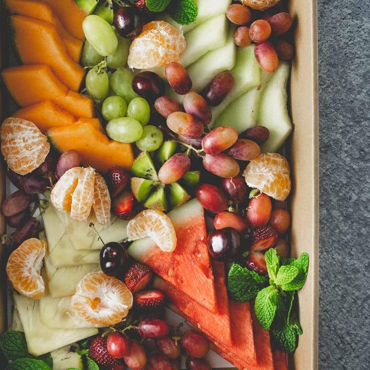 Close up of a box of sliced seasonal fresh fruit including rockmelon, watermelon, grapes, pineapple, kiwi fruit and strawberries on a marble table.