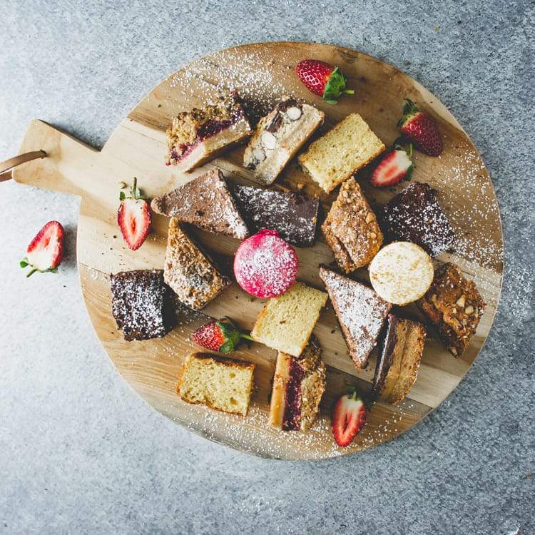 A round wooden board filled with assorted sweet slices and macarons, dusted in icing sugar with a strawberry garnish on a marble table.