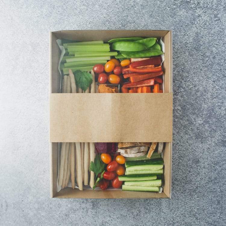 A rectangular box with a clear lid containing three dips, batons of celery, carrot, cucumber, red and green pepper, cherry tomatoes, crostini, and grissini sticks.