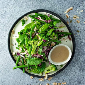 A large round plate with a mix of kale, peas, broccoli, green beans, toasted coconut, mixed seeds, and baby spinach, with a small bowl of red wine viniagrette on a marble table.