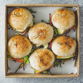 A square cardboard box containing six mini rolls with vegan fillings, such as pumpkin and rocket, eggplant and caramelised onion, or avocado and tomato relish, on a marble table.