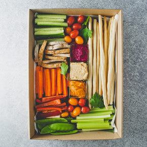 A large rectangular box containing three tubs of dips, batons of celery, carrot, cucumber, red and green pepper, whole cherry tomatoes, crostini, and grissini sticks on a marble table.
